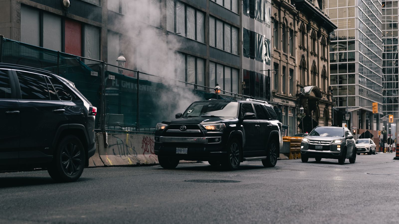 Cars driving through a bustling city street with high-rise buildings and construction barriers.