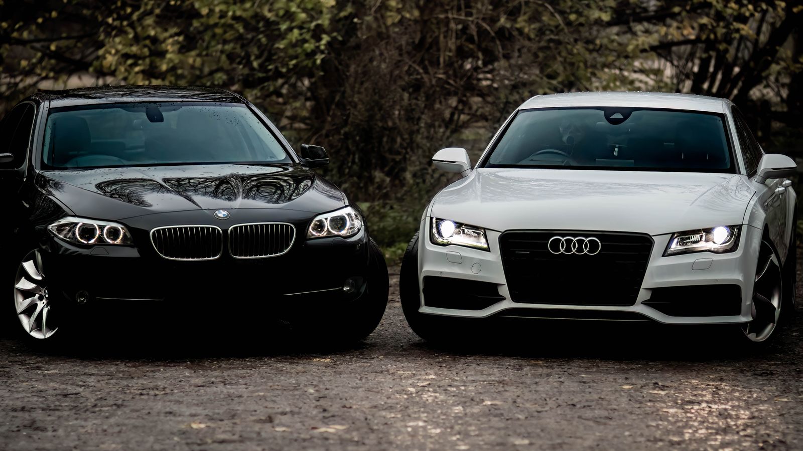 Black BMW and white Audi luxury sedans parked outdoors on a gravel path.