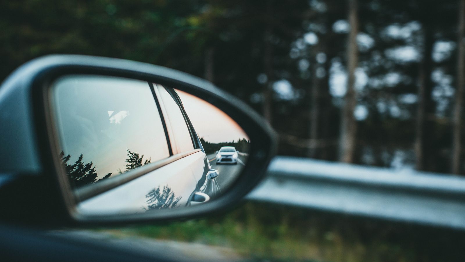 A car is reflected in a side mirror on a scenic forest road.